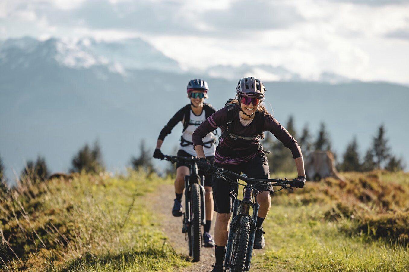 Two cyclists on a mountain trail with a clear sky and distant mountains.