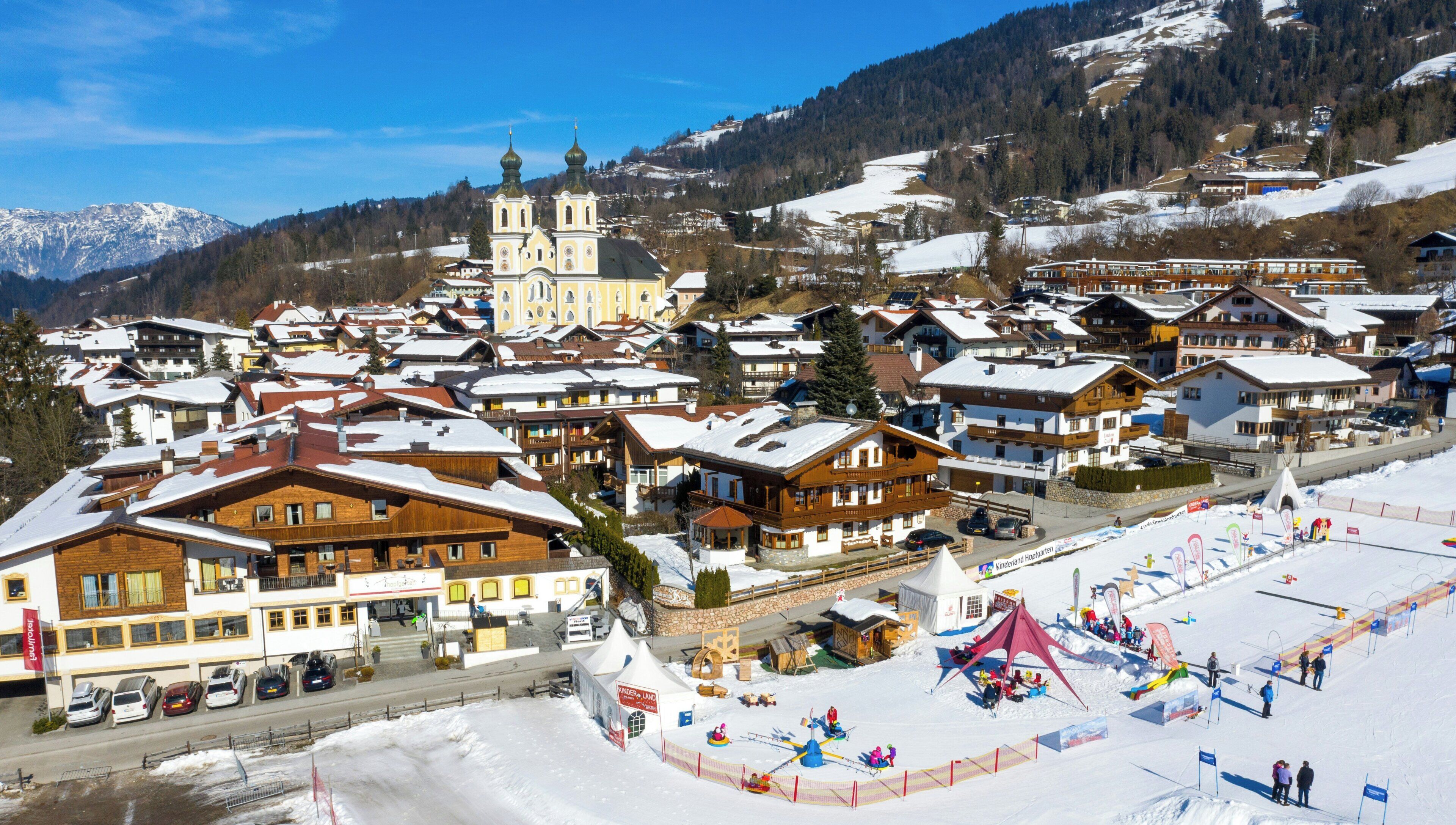 A snow-covered village with traditional wooden houses, a church, and a ski resort area with a tent and flags.