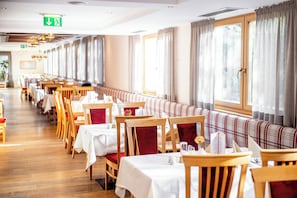 A restaurant with wooden tables and chairs, a long bench seating area, and large windows with curtains. - Das Hopfgarten Aparthotel (Hopfgarten im Brixental)