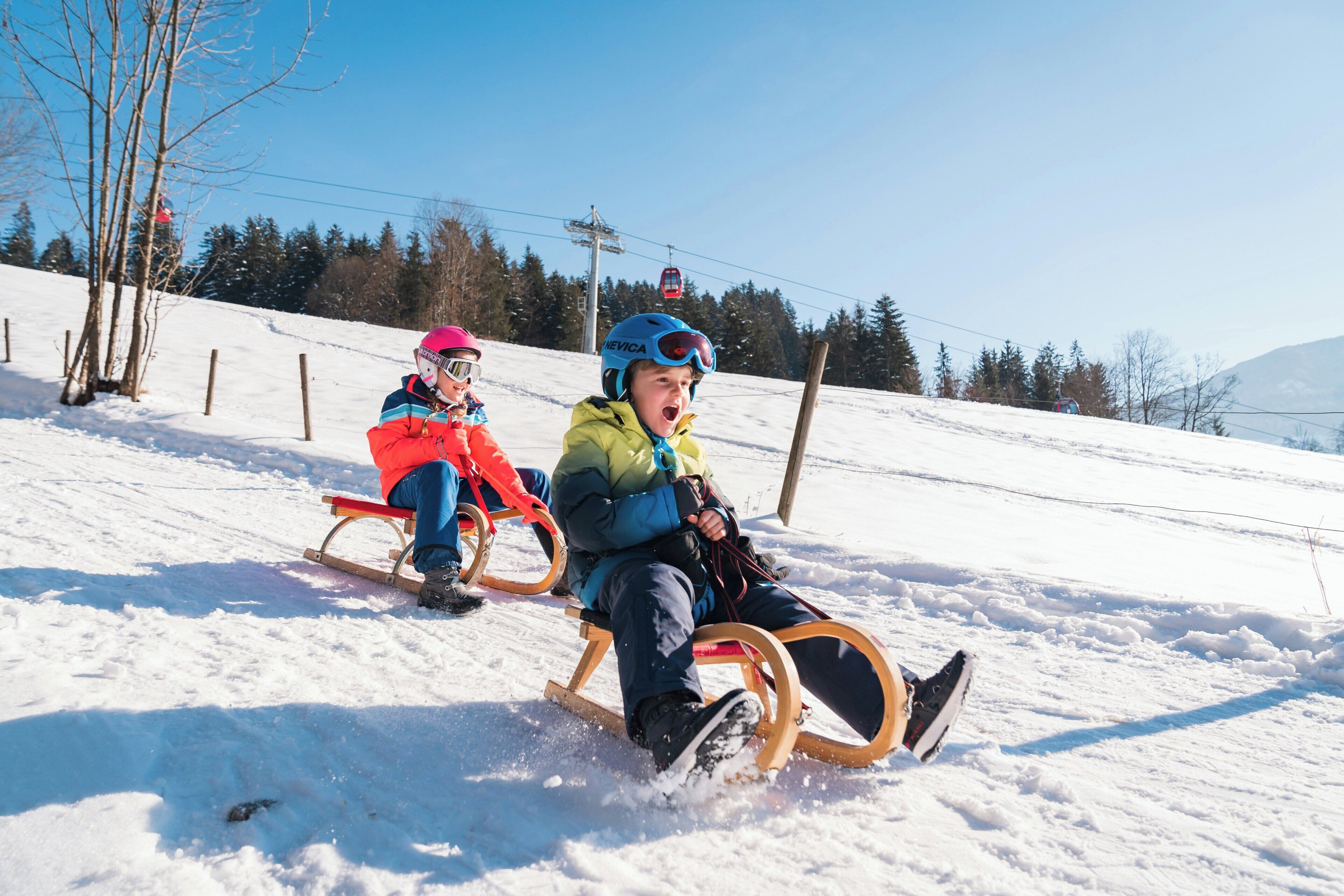 Two individuals sledding on a snowy slope with a clear sky and trees in the background.