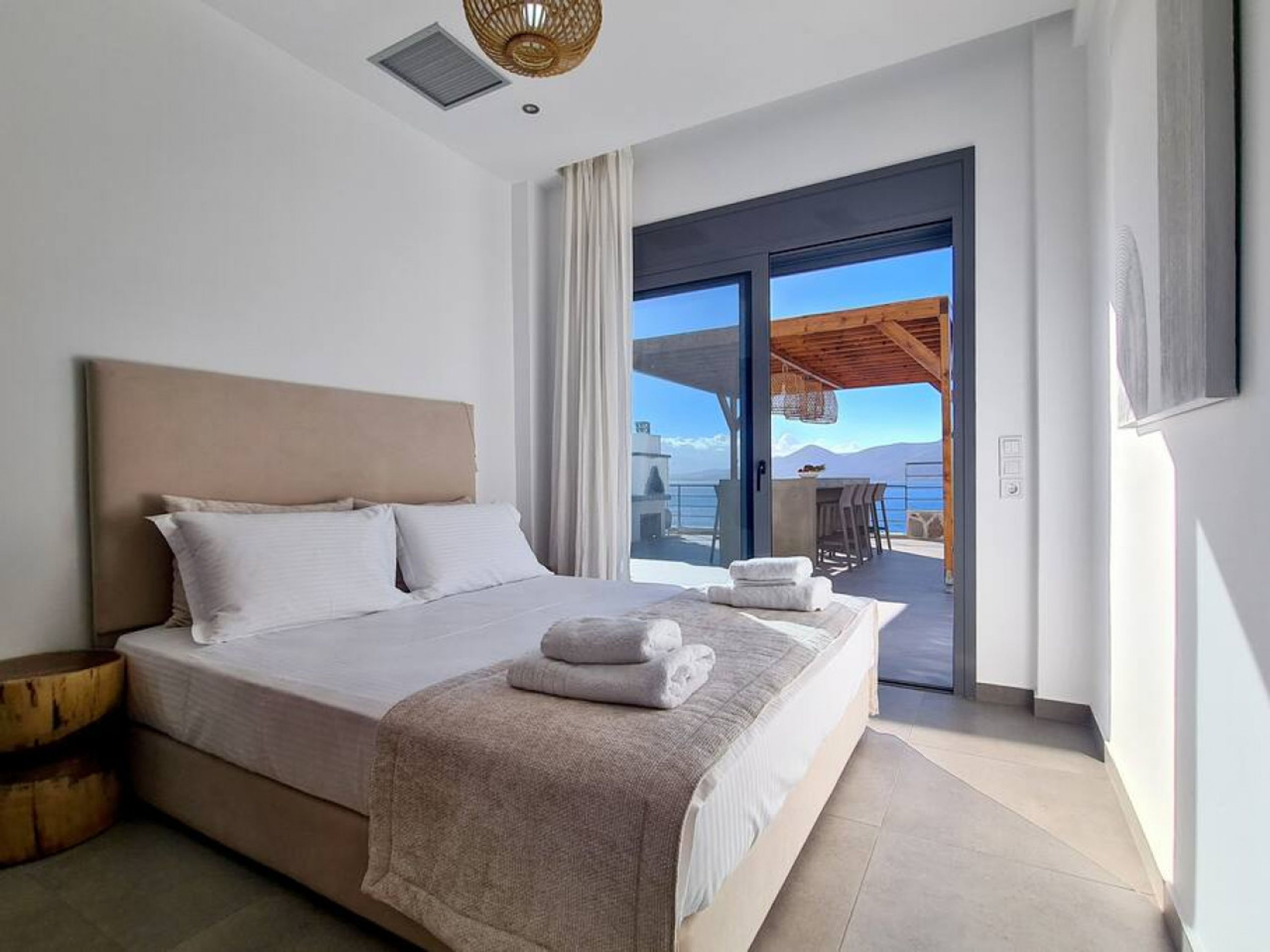 A modern bedroom with a large bed, a wooden side table, and a view of the sea through a sliding glass door.