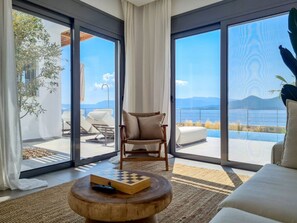 Una sala de estar moderna con un amplio ventanal corredera que da a un área de estar exterior con vista al mar y a las montañas.