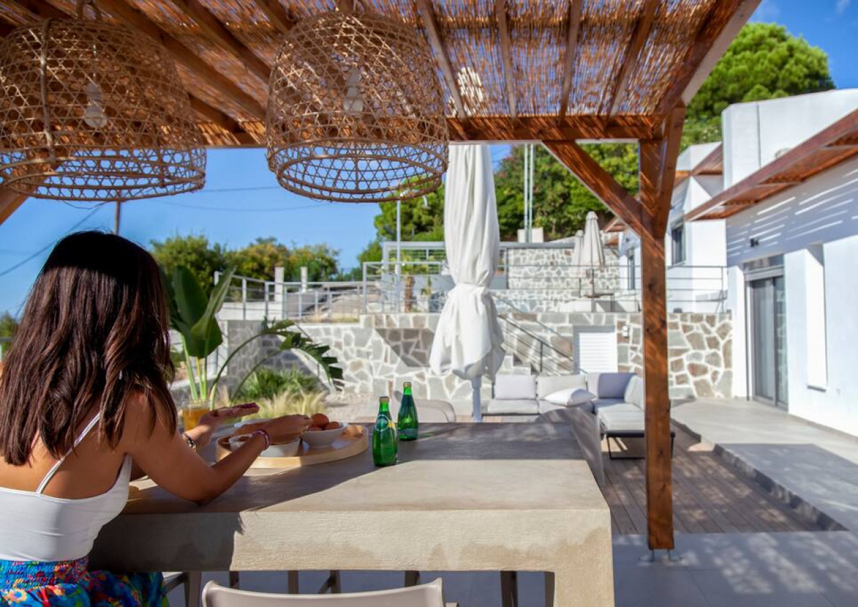 A covered outdoor seating area with a table and chairs, a pergola overhead, and a stone wall in the background.