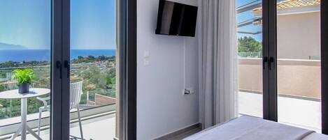 Habitación de hotel con cama, televisor montado en la pared, balcón con mesa y sillas, y vista al mar y al paisaje.