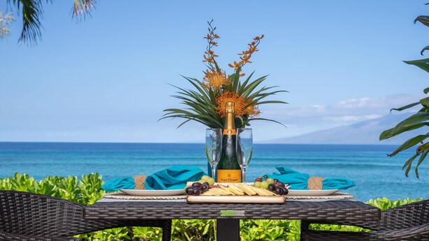 A table set with a bottle of Veuve Clicquot champagne, a glass, and a plate of cheese and grapes, overlooking a scenic ocean view. - Kuleana 405 Oceanfront 1Bedroom 1 Condo by RedAwning (Lahaina)