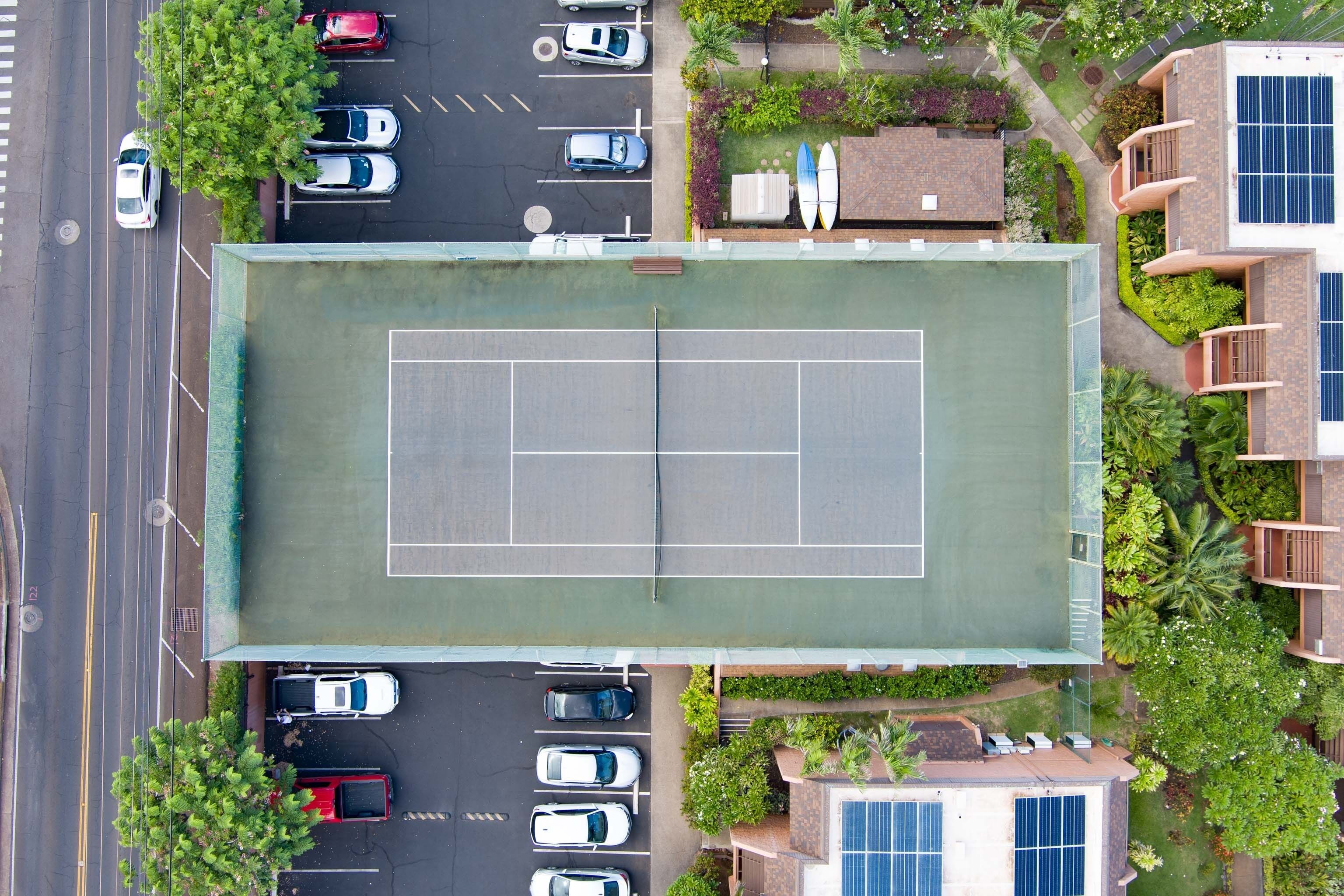 An aerial view of a tennis court surrounded by parked cars, a road, and residential buildings with solar panels.