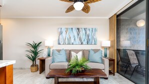 A modern living room with a wooden coffee table, a sofa with blue cushions, a large abstract painting, and a ceiling fan. - Kuleana 405 Oceanfront 1Bedroom 1 Condo by RedAwning (Lahaina)