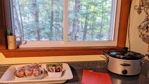 Una cucina con vista su una foresta attraverso una finestra, una pentola a cottura lenta, un vassoio con cibo confezionato e un libretto rosso sul bancone.