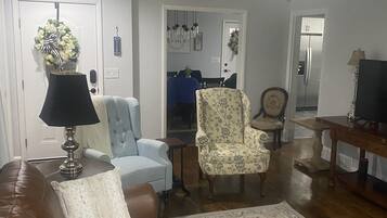 Una sala de estar con un sofá de cuero marrón, una mesa de centro de madera, un sillón floral y un comedor con mantel azul y silla floral.