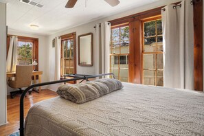 A bedroom with a bed, a desk, and a chair. There are windows with curtains, a ceiling fan, and a view of the outside. - 11th Street River House | Walk to Town | On River (Bandera)