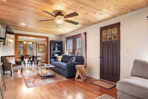 A cozy living room with a dark wooden door, a ceiling fan, and a dining area with a table and chairs. - 11th Street River House | Walk to Town | On River (Bandera)