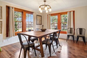 Comedor con mesa de madera, seis sillas, una araña y ventanas grandes con cortinas.