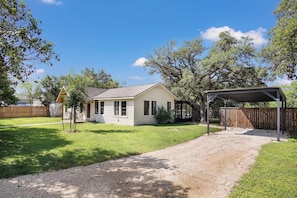 A single-story house with a covered porch, a fenced yard, and a gravel driveway. - 11th Street River House | Walk to Town | On River (Bandera)