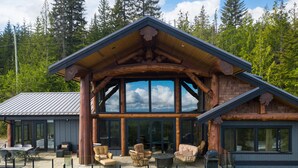 A log cabin with a large glass front, outdoor seating area, and a view of the forest.