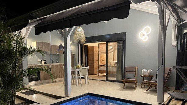 Un espace piscine extérieur moderne, doté d’un coin repas à l’abri derrière des parois de verre et d’une cuisine visible à travers des portes vitrées.