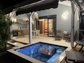 Un espace piscine extérieur moderne, doté d’un coin repas à l’abri derrière des parois de verre et d’une cuisine visible à travers des portes vitrées.