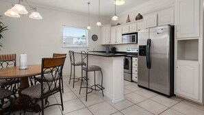 A kitchen with white cabinets, a stainless steel refrigerator, a microwave, and a dining area with a wooden table and metal chairs.