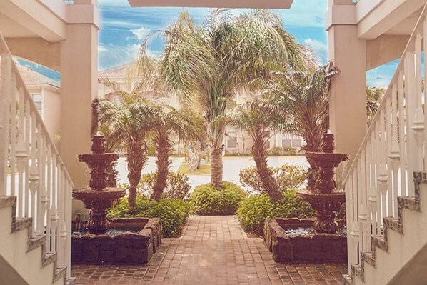 A covered walkway with a brick floor, flanked by white railings and palm trees.