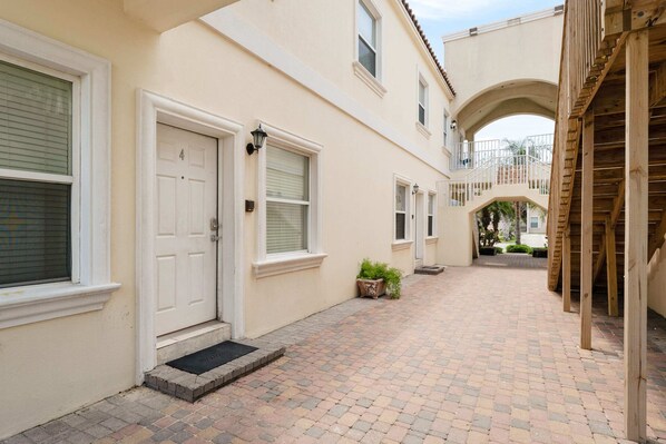 Un patio estrecho y empedrado con una puerta blanca y ventanas, que da paso a una escalera de madera.