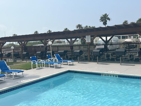 A swimming pool with blue-tiled edges, surrounded by blue lounge chairs and barbecue grills under a wooden pergola.