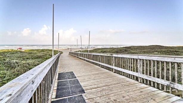 A wooden boardwalk leading to a beach with a grassy area on one side and a wooden fence on the other.