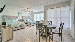 A bedroom with two beds, a dining table with chairs, a ceiling fan, and a large window with blinds.