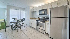 A compact kitchen with white cabinets, a stainless steel refrigerator, a microwave, and an oven. There is a dining table with chairs and a window with blinds.