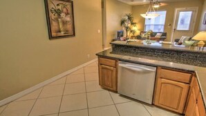 A kitchen with a granite countertop, wooden cabinets, and a dishwasher. There is a painting on the wall and a window with blinds.