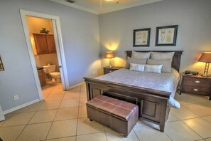 Un dormitorio con una cama grande, una mesita de noche, un banco y un baño visible a través de una puerta abierta.