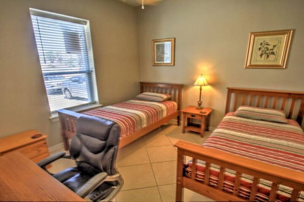A bedroom with two beds, a desk, a chair, a lamp, and a window with blinds.