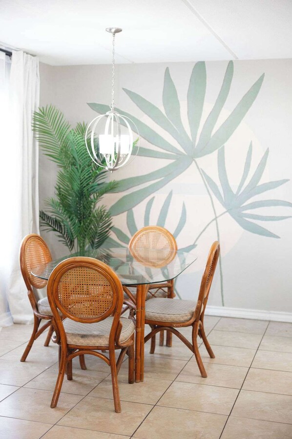 A dining area with a glass table surrounded by wicker chairs, a hanging pendant light, and a large plant with broad leaves.