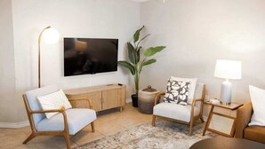 Una sala de estar con un televisor de pantalla plana montado en la pared, un mueble de madera, dos sillas de mimbre, una lámpara de pie, una mesita de noche con lámpara y una planta grande en maceta.