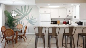 Una cocina moderna con armarios blancos, una isla central con taburetes y un comedor con sillas de mimbre.