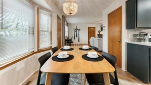 Een moderne keuken met een eethoek, met een houten tafel met vier stoelen, een kroonluchter en uitzicht op buiten door grote ramen.