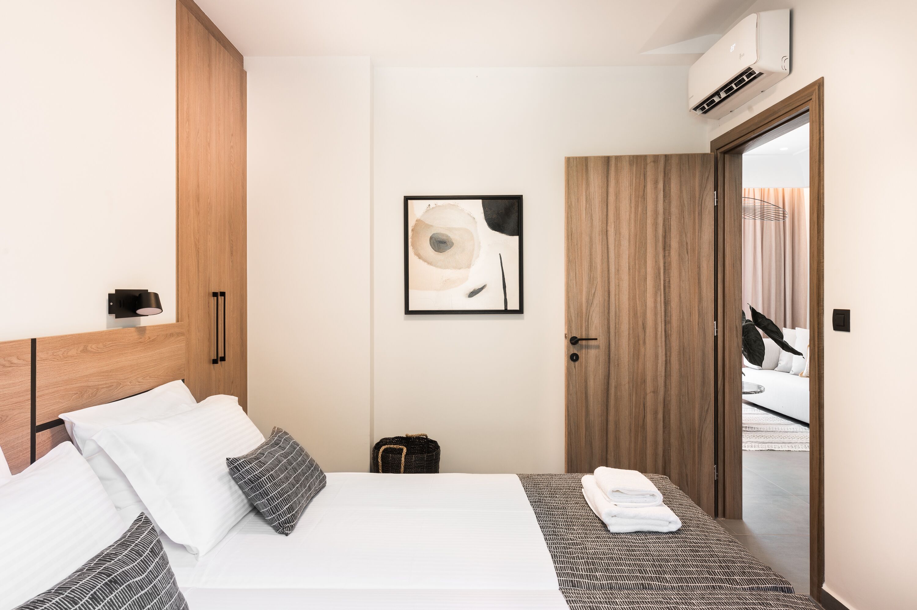 A modern hotel room with a wooden headboard, a bed with white and patterned bedding, a small bedside table with a lamp, and a framed abstract artwork on the wall.