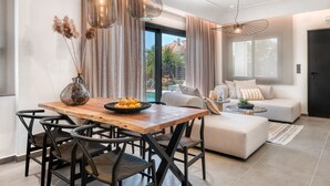 Un salon moderne avec un canapé, une table à manger et des chaises. On y trouve une grande fenêtre avec des rideaux, un luminaire suspendu et une porte à l’arrière-plan.