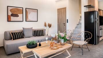 Una sala de estar moderna con sofá, una mesa de centro y objetos decorativos. Una escalera conduce a una zona de cocina con un refrigerador.