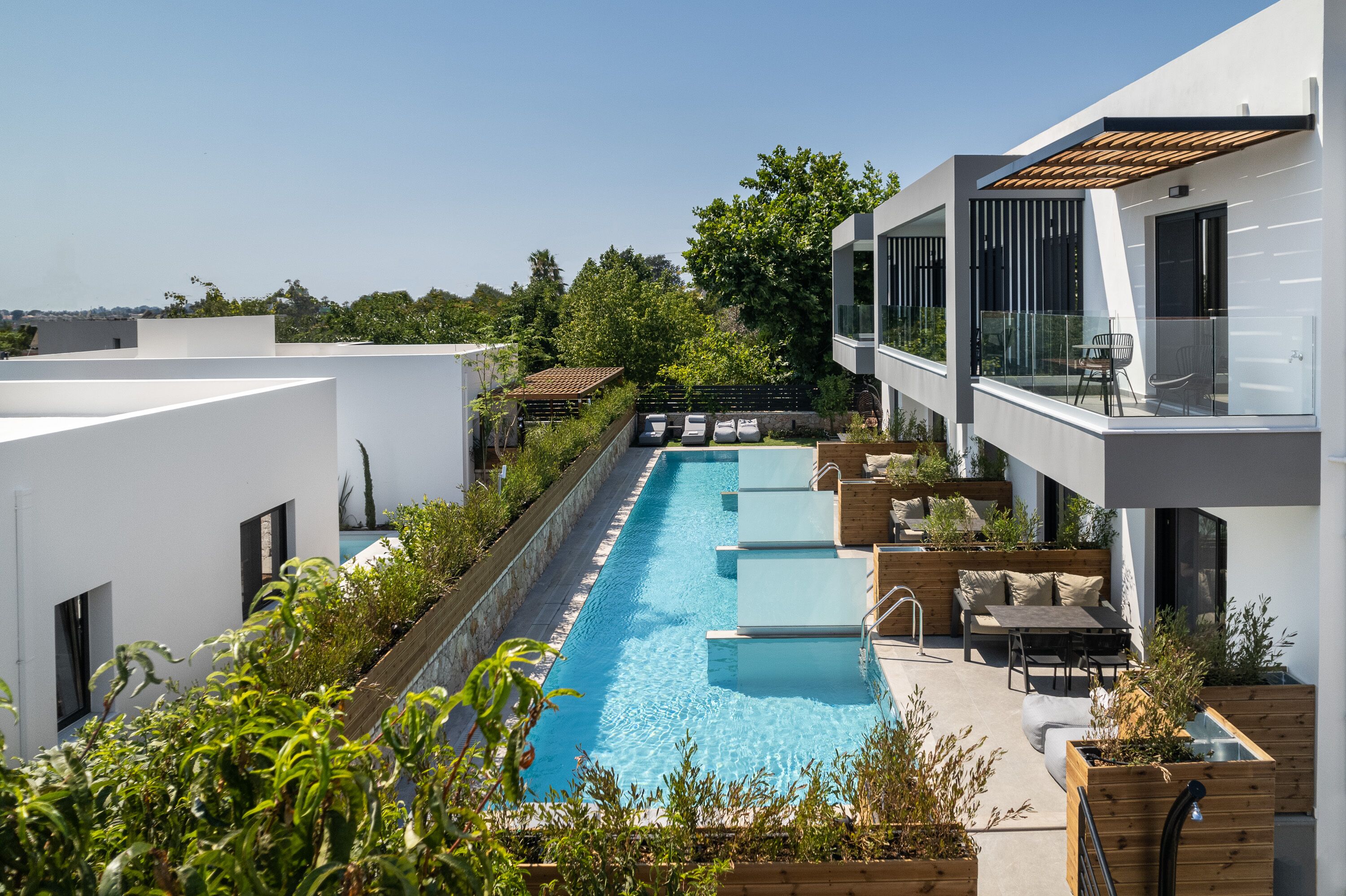 Un área de piscina moderna con una piscina rectangular larga, rodeada de asientos y jardineras de madera, con vistas a un jardín exuberante y un cielo despejado.