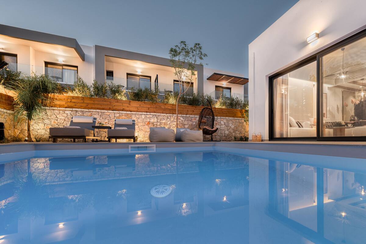 Un área de piscina moderna al aire libre con un muro de piedra, sillas de descanso y un gran ventanal que da a una sala de estar.