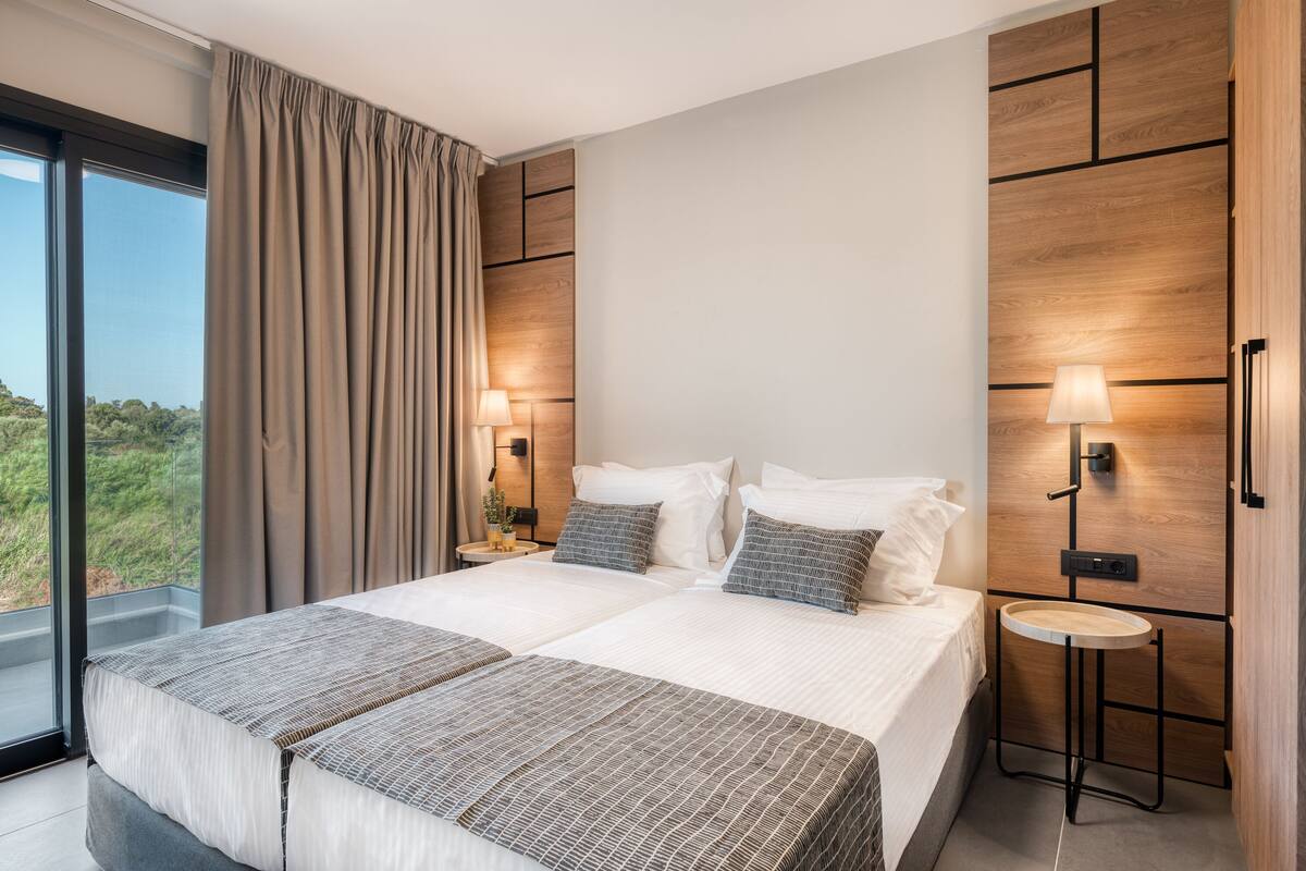 Habitación de hotel moderna con una cama grande, dos mesitas de noche, una pequeña mesa redonda y vistas al exterior.