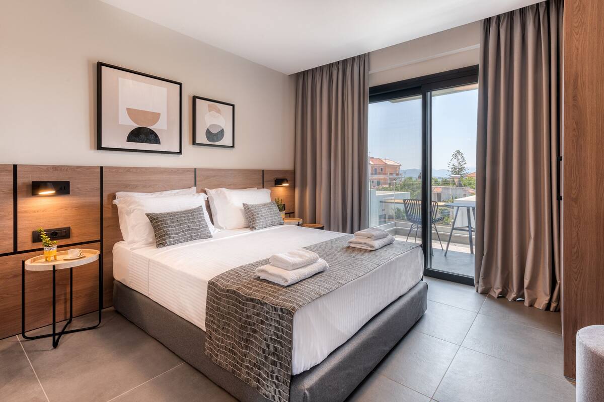 Una habitación de hotel moderna con una cama grande, una mesita pequeña, una mesita de noche y vistas al exterior a través de un ventanal.