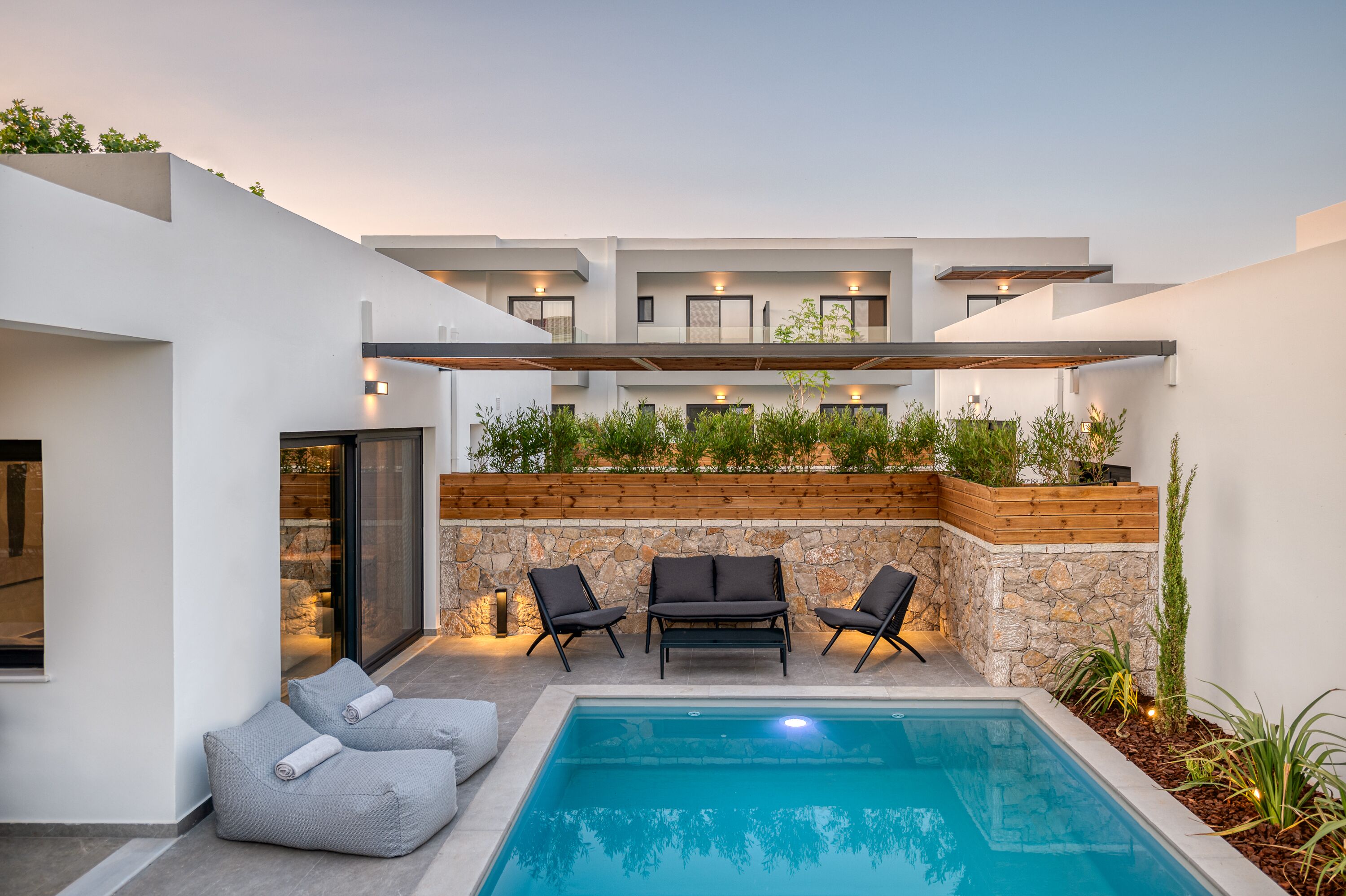 Un'area piscina moderna all'aperto con muro in pietra, terrazza in legno e poltrone relax nere.