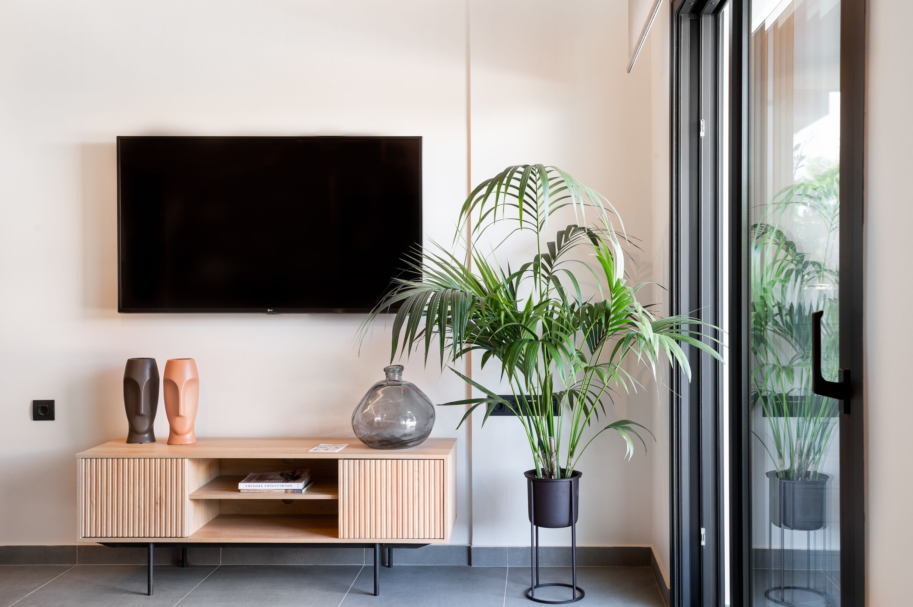 A modern living room with a flat-screen TV mounted on the wall, a wooden console table with decorative vases, and a potted plant.