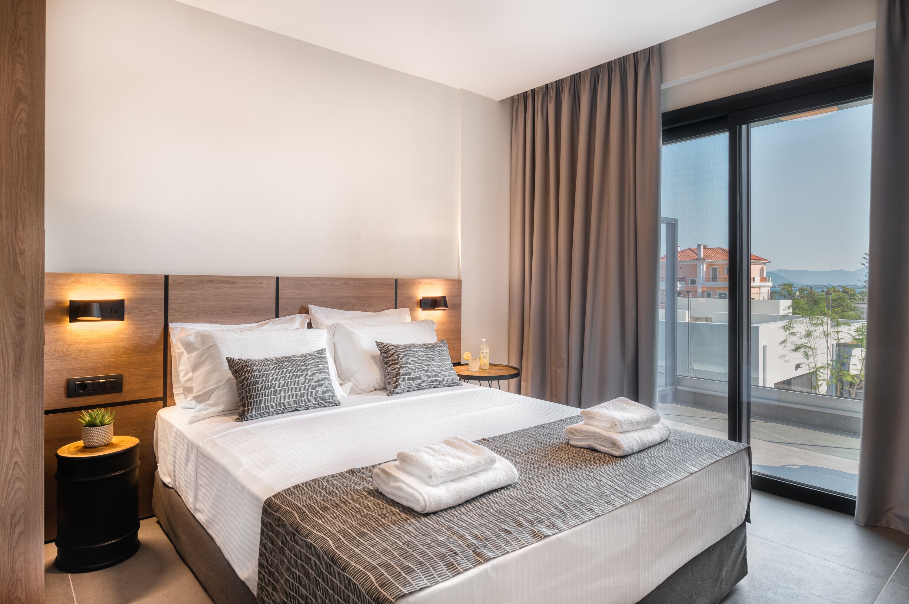 Ein modernes Hotelzimmer mit einem großen Bett, einem Nachttisch mit Lampe und Blick auf die Stadt durch ein großes Fenster.