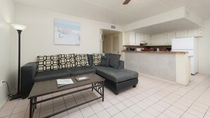 A living room with a sectional sofa, a coffee table, a ceiling fan, and a kitchen area with cabinets and a refrigerator. - Lovely 2 Bed 2 Bath 2nd Floor Condo By Beach (South Padre Island)