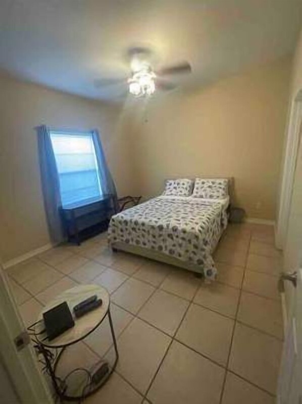 A bedroom with a bed, a ceiling fan, a window with curtains, and a small table with a laptop.