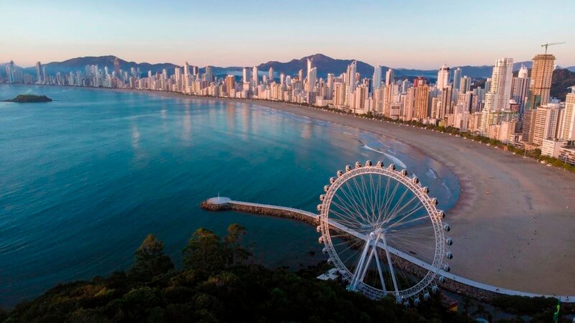 A coastal cityscape with a prominent Ferris wheel, high-rise buildings, and a clear view of the ocean.
