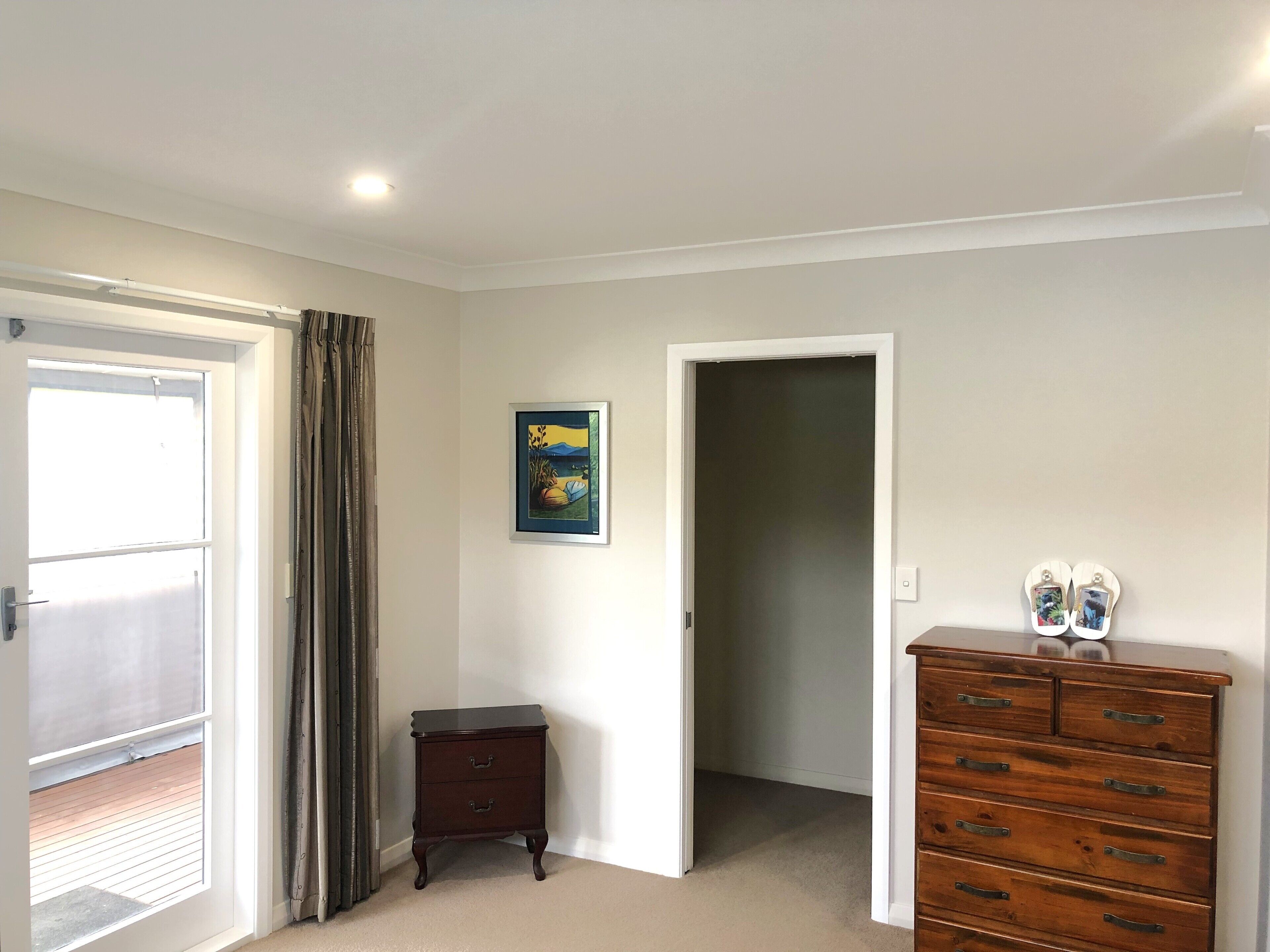 A room with a sliding glass door, a small wooden nightstand, a dresser with drawers, a framed picture on the wall, and a carpeted floor.