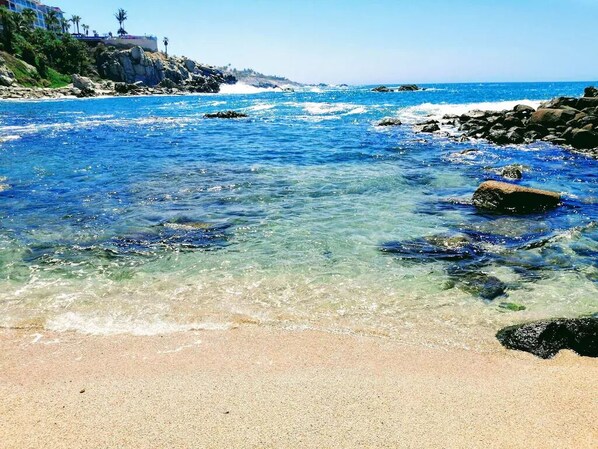 A beach with clear blue water, rocky shoreline, and a distant building on a hill. - REMODELED UNOBSTRUCTED OCEANVIEW GROUND FLOOR 1 BEDROOM All US/CA TV CHANNELS (Cabo San Lucas)