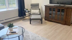 Ein Wohnzimmer mit einem grauen Sessel, einem Glastisch, einem Holzschrank mit Fernseher und einem großen Fenster mit Vorhängen.